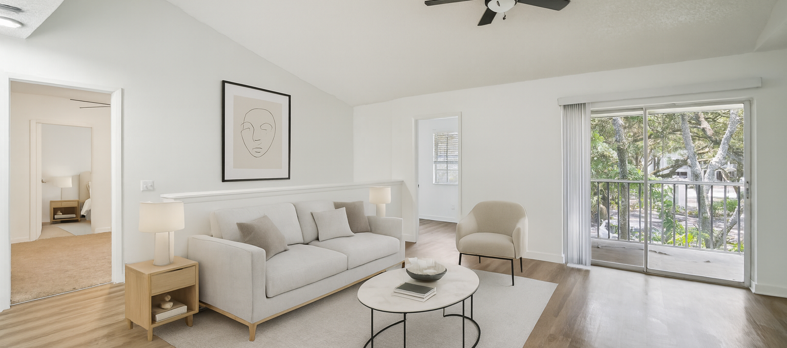 A serene and modern living room features a plush, light-colored sofa adorned with throw pillows, positioned centrally on a large area rug. The room is accented with a contemporary coffee table holding a few decorative items, and a stylish side table with a lamp. A comfortable armchair sits nearby, and a large framed artwork hangs above the sofa. The space is brightened by natural light streaming through a sliding glass door leading to a balcony, creating a tranquil atmosphere perfect for relaxation. The room's minimalist design and neutral color palette enhance its inviting and cozy ambiance.